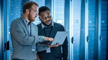 Two technology online graduates in server room looking at analytics on laptop
