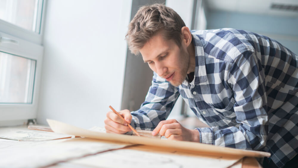 A male construction project manager holding a pencil stands at a drafting table examining a large construction plan.