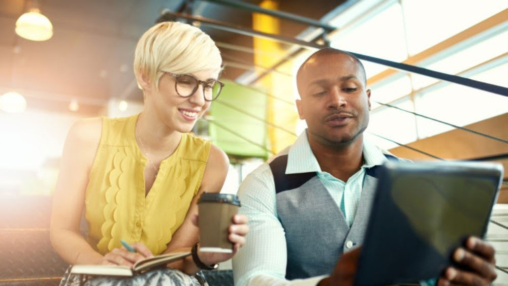 Two social media managers sit on stairs and drink coffee while brainstorming strategy on a tablet.