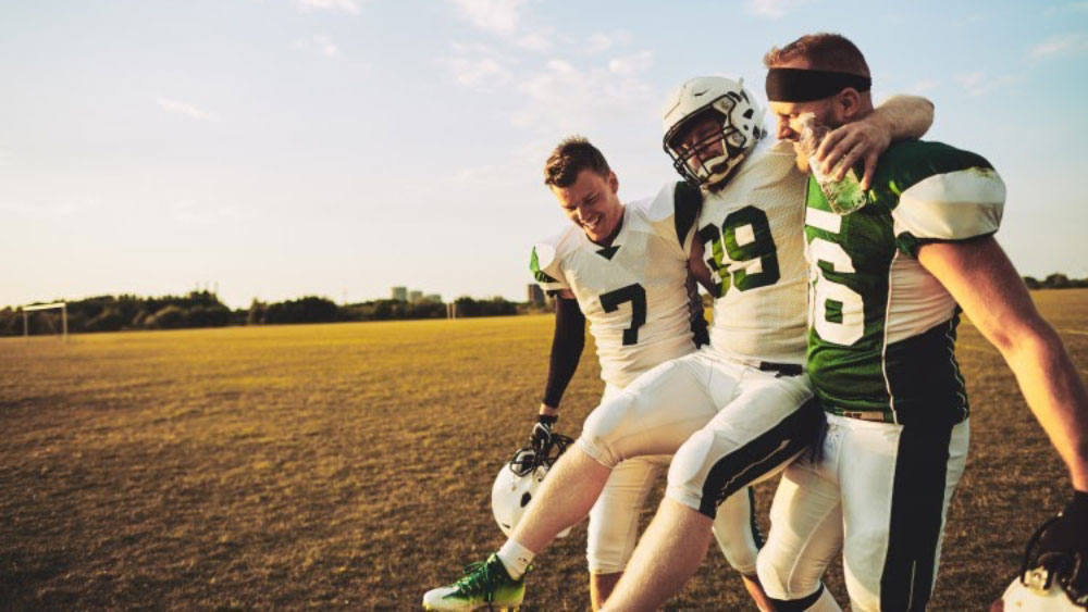 Two football players carry injured teammate across a field.